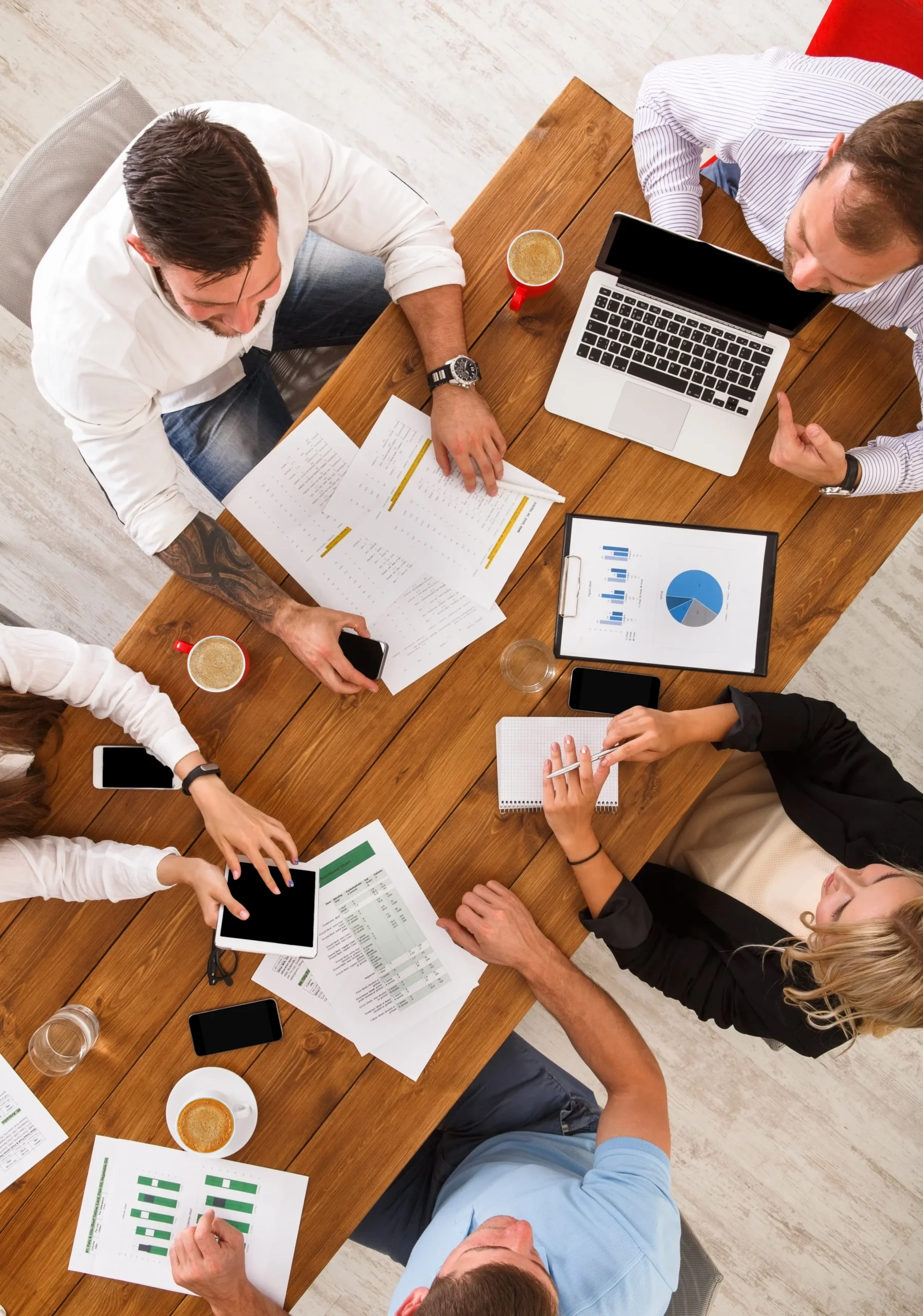 Aerial view of a business team in a strategic planning session, viewed from above a wooden table. Five professionals, three men and two women, are deeply engaged in a discussion over laptops, tablets, and documents, focusing on optimizing their voice communication technologies. Their collaborative efforts are centered around a laptop showing vibrant charts, illustrating their commitment to enhancing user experience and system efficiency.